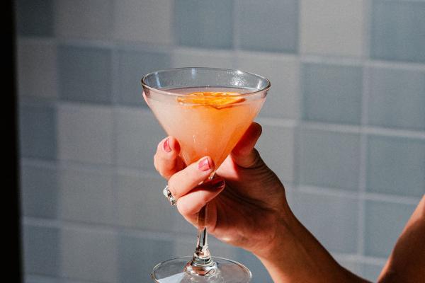 bright orange cocktail in a martini glass being held by a woman's hand in front of a light blue textured wall