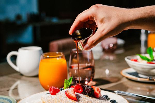 View of large pancake stack topped with strawberries and blueberries with a person'a hand in frame, pouring syrup out of small container onto the top of the pancakes
