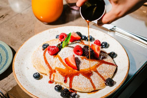 A hand pouring syrup out of a small sauce container onto the top of a stack of pancakes garnished with strawberries and blueberries.