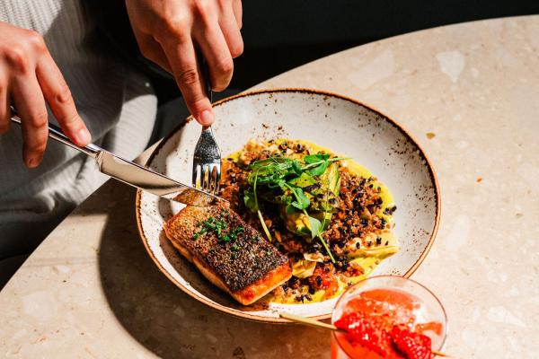 man holding a fork and knife while cutting into a large plate of salmon and quinoa with a bright red cocktail also placed on table