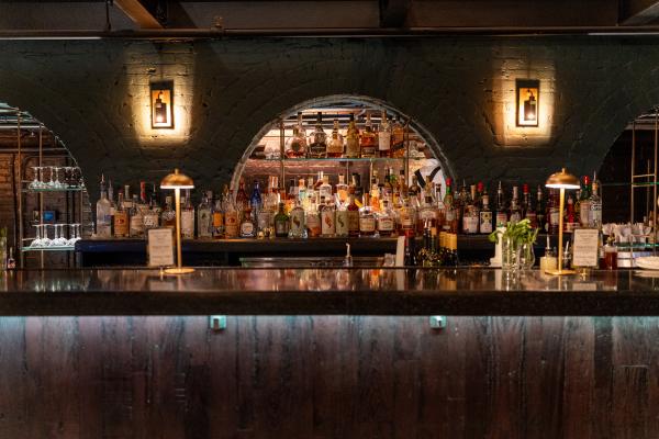 District Still's cocktail bar set within a dark arched brick alcove, lit by wall sconces and lined with glassware and wine bottles.