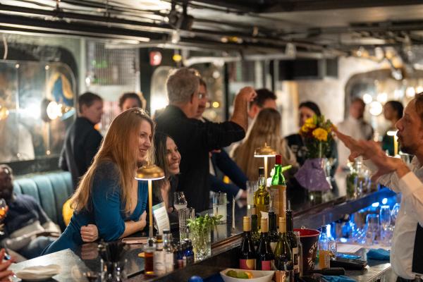 Guests mingling and being served at a crowded bar during a packed evening event, framed by exposed pipes and glowing blue accent lighting.