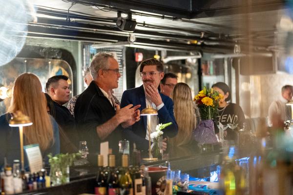 A lively crowd gathered along a busy bar during an event, with bartenders mixing drinks amid bottles, glassware, and warm industrial lighting.